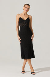 A woman is wearing a black lace midi dress with spaghetti straps, featuring a fitted bodice with a sweetheart neckline and a sheer lace panel at the waist. The skirt falls below the knees with a scalloped hem, and she pairs the dress with black open-toe heels.